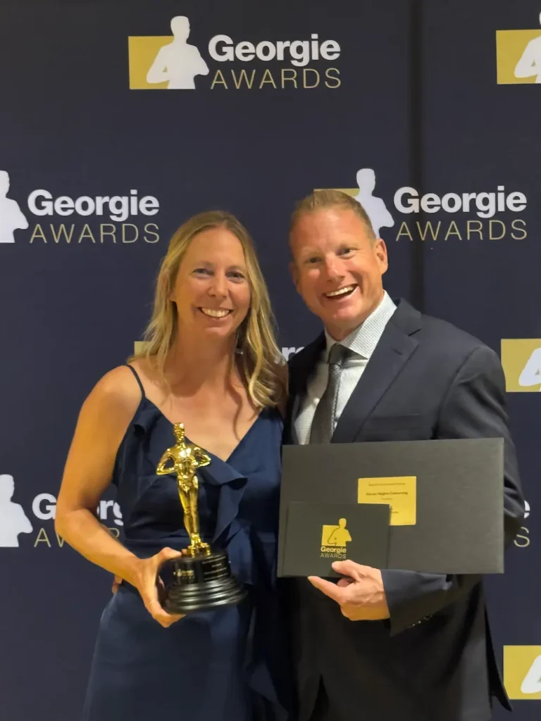 A smiling woman in a blue dress holds a trophy, and a smiling man in a suit holds a certificate as they celebrate their 2026 win at the Georgie Awards, standing proudly in front of the Perch-sponsored backdrop.