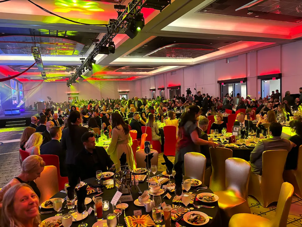 A large, elegant banquet hall filled with people seated at round tables set for dinner. The room is decorated with red and gold lighting for the 2026 Georgie Awards, and there is a stage at the front with bright spotlights.