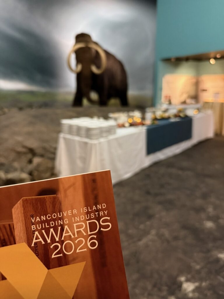 A brochure for the Vancouver Island Building Industry (VIBI) Awards 2026 is held in the foreground, with a buffet table and a large woolly mammoth display in the background—perfect inspiration for any custom home builder from Falcon Heights.