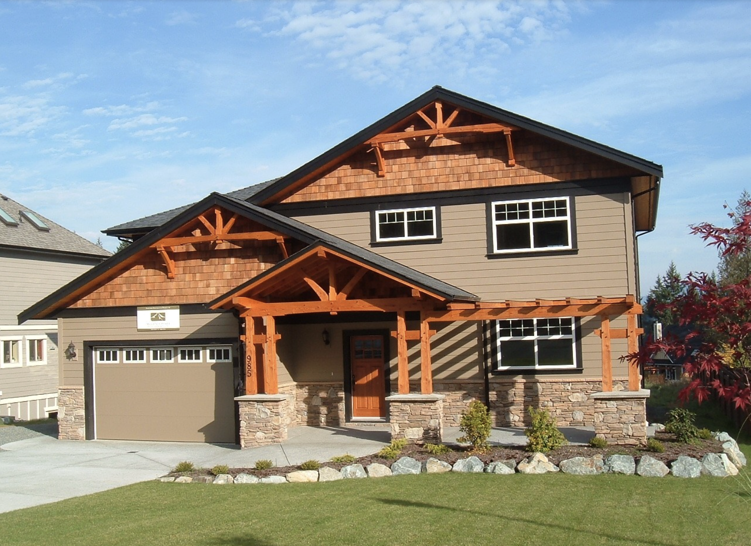 Two-story Craftsman-style house with tan siding, stone accents, and wood trim. Located on Vancouver Island, this high-performance homebuilding features a covered front porch, attached garage, manicured lawn, and a clear blue sky in the background.