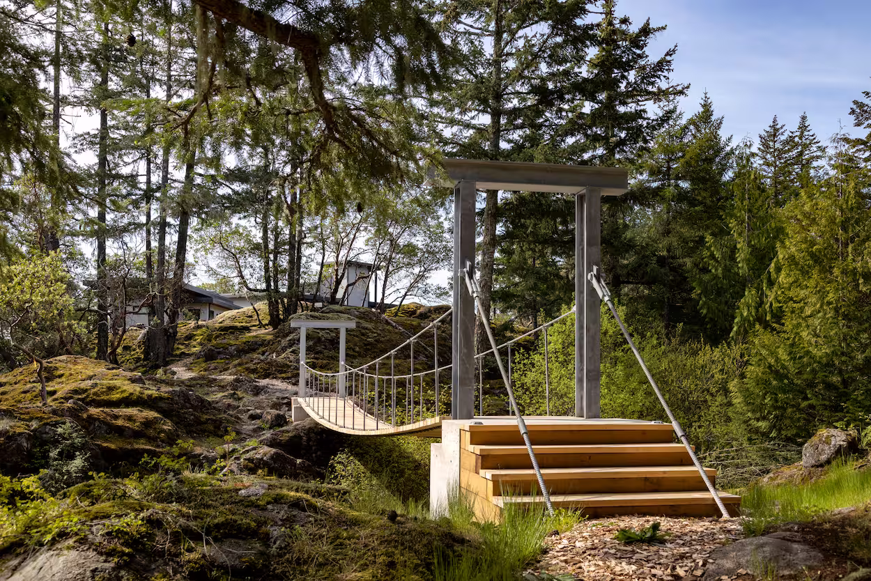 A small wooden suspension bridge with metal cables spans a mossy, rocky landscape surrounded by tall trees in Falcon Heights, with a custom home builder's house partially visible in the background under a clear sky.