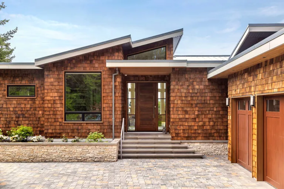 Modern house exterior with cedar shingle siding, large windows, a wooden front door with glass panels, stone steps, a brick walkway, and a landscaped front garden bed with small plants.