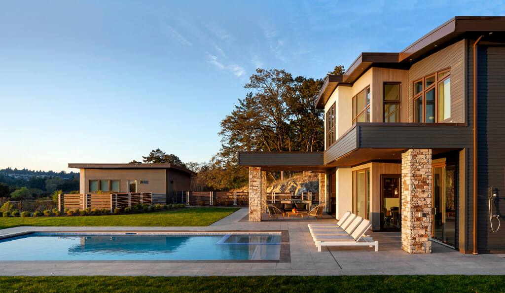 Modern two-story house with large windows, stone accents, and patio seating. A swimming pool with clear blue water sits in the backyard, surrounded by manicured lawn and distant trees.
