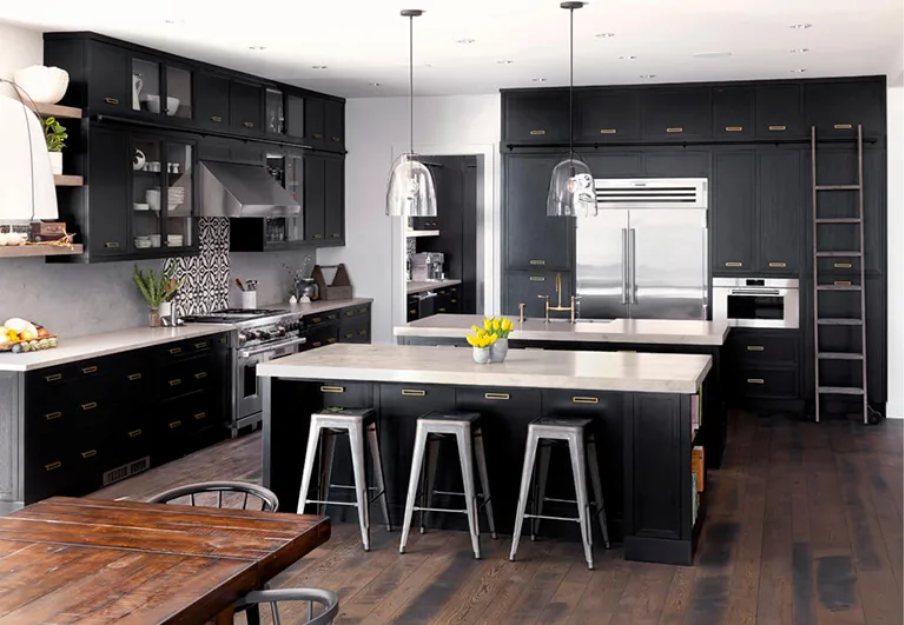 Modern kitchen with black cabinets, two islands with white countertops, stainless steel appliances, pendant lights, a ladder on the right, and wooden floors. A bowl of lemons sits on one island. Dining table is in the foreground.