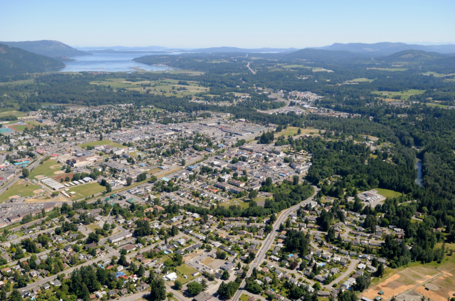 Aerial view of a small town surrounded by green hills, forests, and distant bodies of water under a clear blue sky. Residential and commercial areas are visible along winding roads.