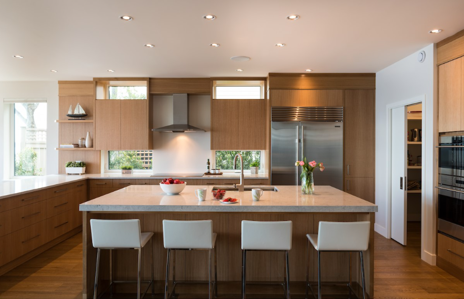 A modern kitchen with wood cabinets, a large central island featuring four white bar stools, and a stainless steel refrigerator, all captured in a warm lighting scene.